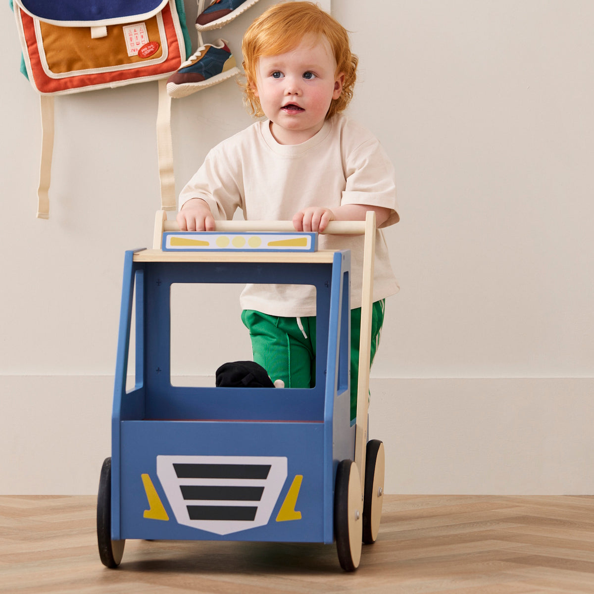 Front view of a toddler with reddish hair holding onto the wooden handle of a blue toy walker truck.