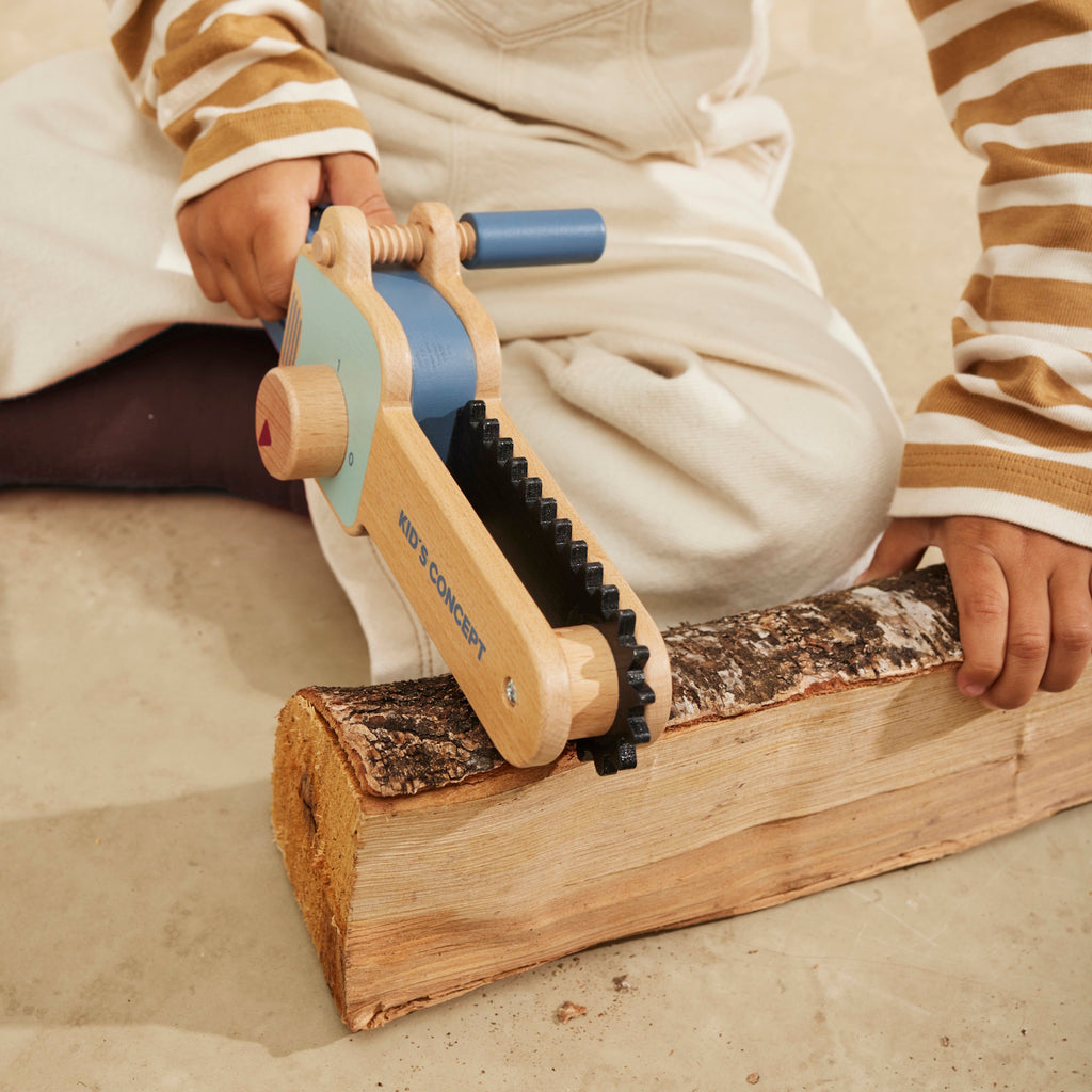 Wooden toy chainsaw with blue handle and details, featuring a rotating knob and black ridged edge. "Kid’s Concept" is printed on the side.