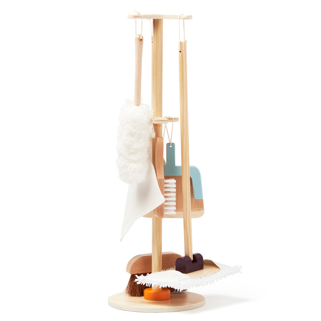 A wooden toy cleaning set on a stand includes a mop, broom, fluffy duster, and scrub brush. The set conveys a playful and organized tone.