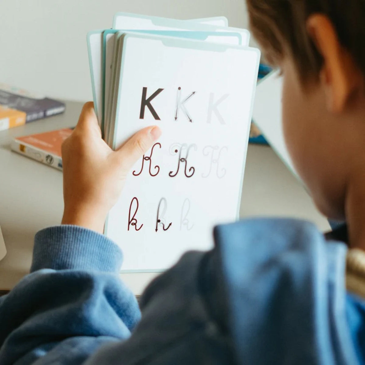 A child's hand holds a white educational card showing uppercase and lowercase 'K' tracing guides in print and cursive.