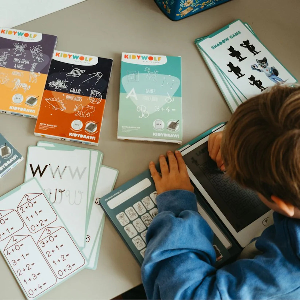 A child uses a stylus on a Kidywolf drawing tablet surrounded by educational activity cards for math, letters, and games.
