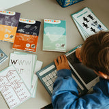 A child uses a stylus on a Kidywolf drawing tablet surrounded by educational activity cards for math, letters, and games.