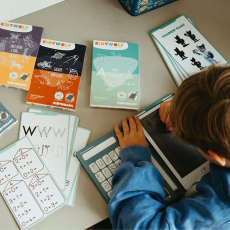 A child uses a stylus on a Kidywolf drawing tablet surrounded by educational activity cards for math, letters, and games.
