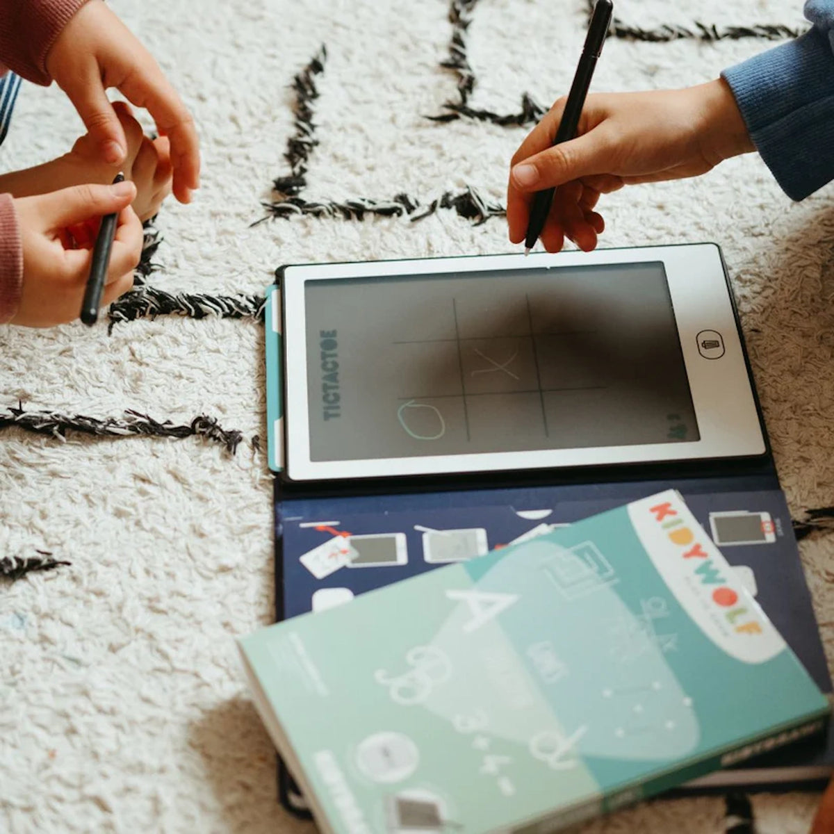 Two children playing tic-tac-toe on a Kidywolf LCD writing tablet with pens on a white shag rug.