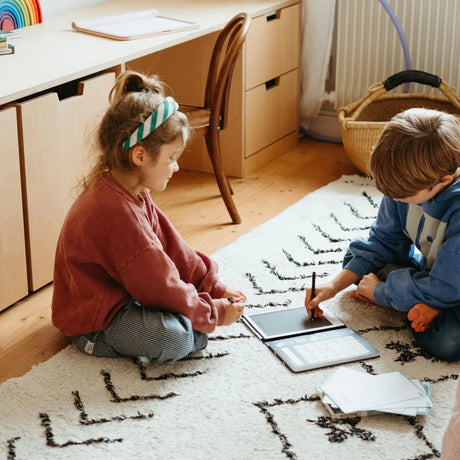 Two children sitting on a rug using a Kidydraw Mini tracing tablet for educational drawing and writing games.