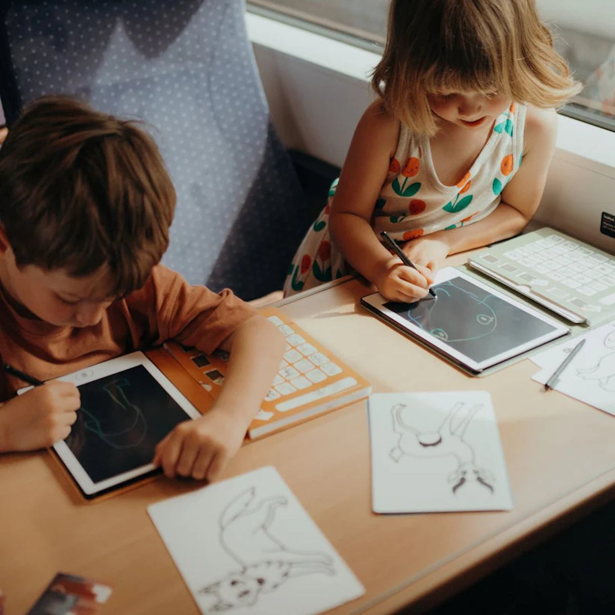 Two children use styluses to draw on Kidywolf LCD tablets while sitting at a table during a train or bus ride.