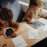 Two children use styluses to draw on Kidywolf LCD tablets while sitting at a table during a train or bus ride.