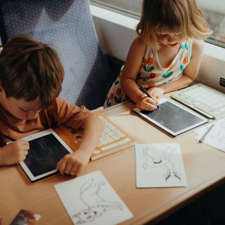Two children use styluses to draw on Kidywolf LCD tablets while sitting at a table during a train or bus ride.