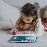 A young girl lying on a couch and drawing with a pencil on an illuminated tracing pad kit.