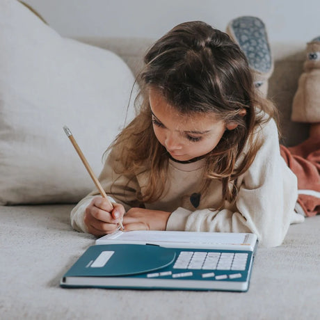 A young girl lying on a couch and drawing with a pencil on an illuminated tracing pad kit.