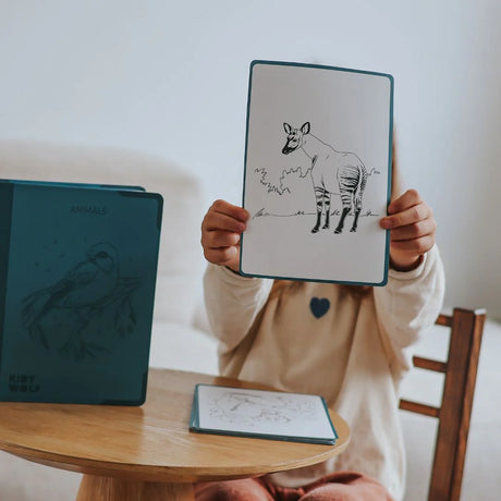 A child holds up a drawing of an okapi in front of their face, next to a drawing kit case.