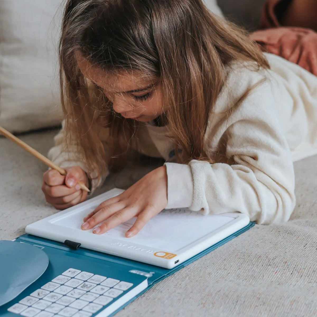 A young girl lying down while drawing on an illuminated tracing pad with a pencil.