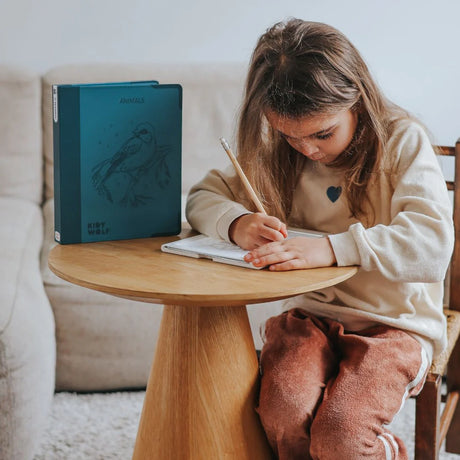 A young girl sits at a wooden table, focused on drawing with a pencil on an illuminated sketching pad.