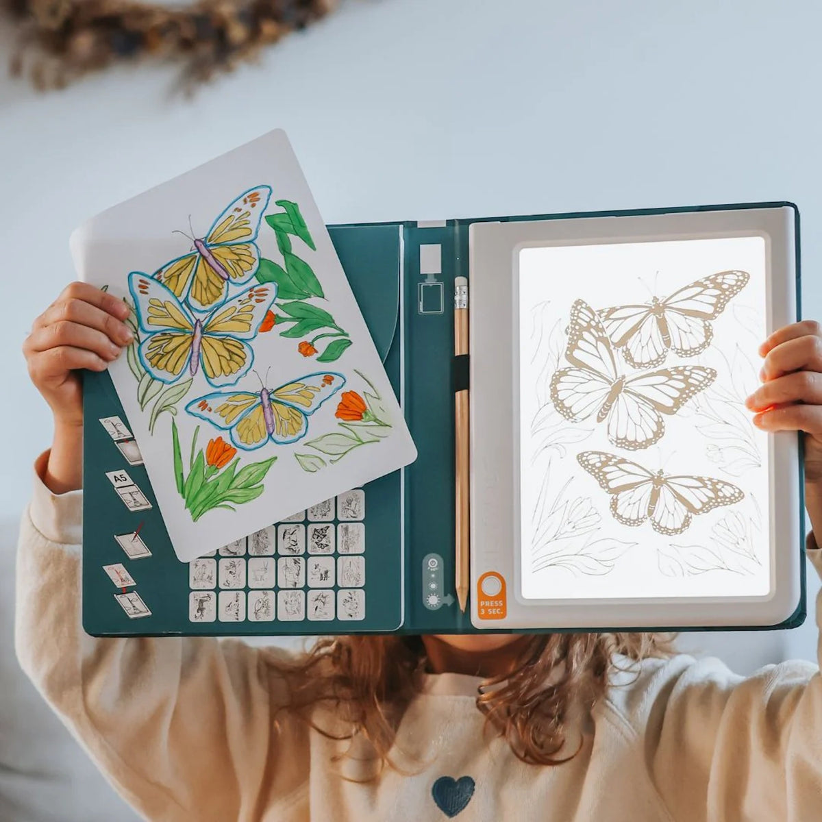 A child holds up a lit drawing pad with a butterfly sketch and a colored drawing of butterflies.