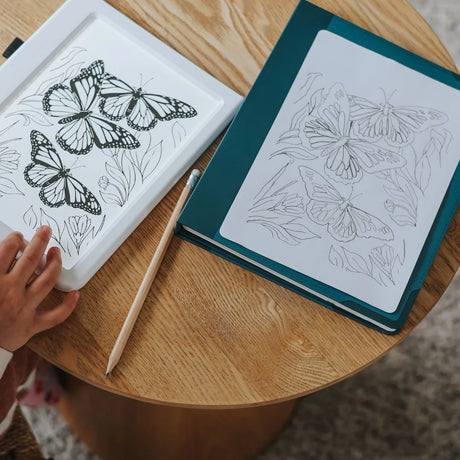 A child's hand near a white light-up pad and a green sketch pad, both showing butterfly drawings on a table.