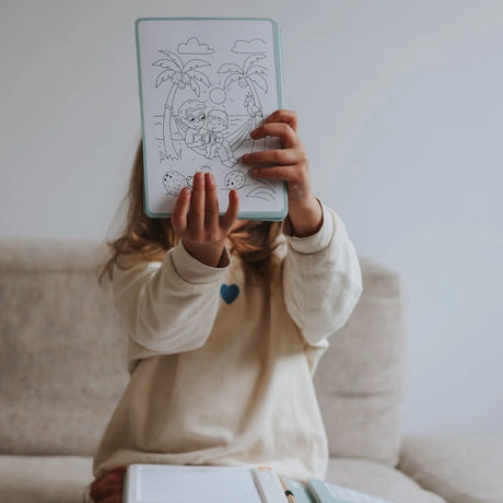 A child holds up a completed tropical beach coloring page, hiding their face while sitting on a couch.