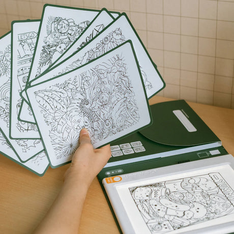 A hand holding a fan of coloring sheets over a green drawing pad folder and an illuminated tracing tablet.