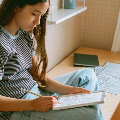 A girl sitting cross-legged draws with a pencil on an illuminated light-up sketching pad in her room.