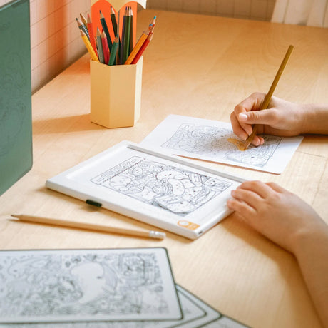 A child coloring a sheet on a wooden table next to an illuminated tracing pad and a cup of colored pencils.