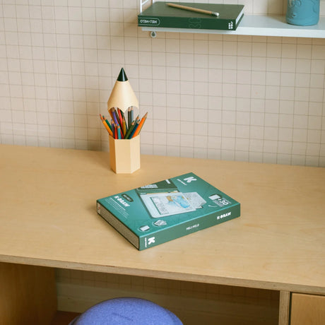 A green K-DRAW box sits on a wooden desk next to a pencil-shaped holder filled with colored pencils.