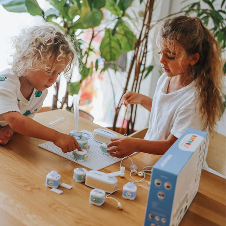 Two young children sitting at a wooden table, playing with an electronic building blocks toy kit.