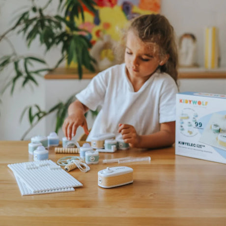 A young girl sits at a wooden table playing with a Kidywolf electronic toy building set.