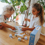 Two young children at a wooden table playing with a modular electronic toy set surrounded by green plants.