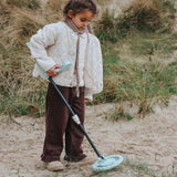 A young child using a light blue toy metal detector on a sandy, grassy beach.