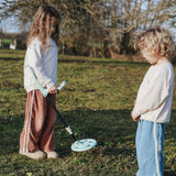 Two children standing in a grassy field, one holding a light blue toy metal detector.