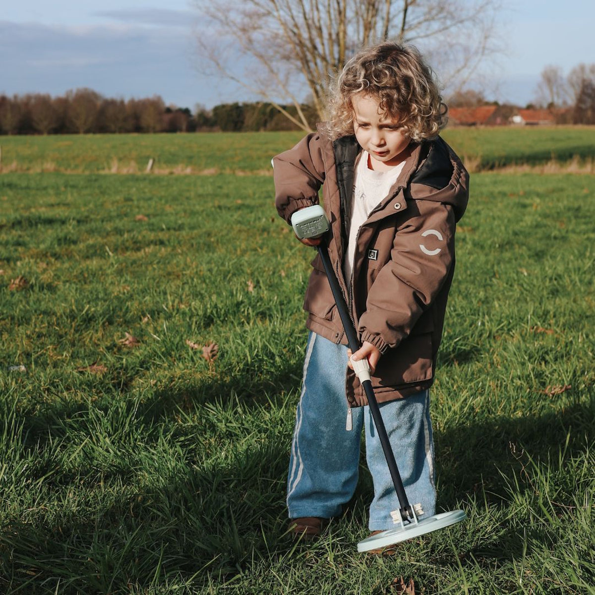 A young child in a brown jacket using a toy metal detector in a grassy green field on a sunny day.