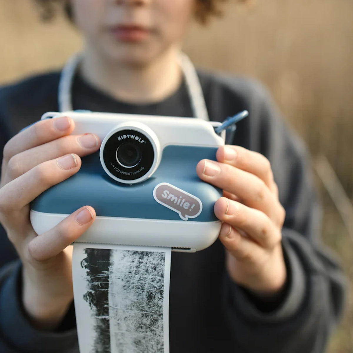 Child holding a blue and white Kidywolf Kidyprint instant camera printing a black and white thermal photo outdoors.