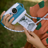 Child holding a blue KIDYWOLF KIDYPRINT camera printing a black and white thermal photo outdoors.