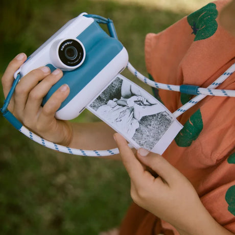 Child holding a blue KIDYWOLF KIDYPRINT camera printing a black and white thermal photo outdoors.