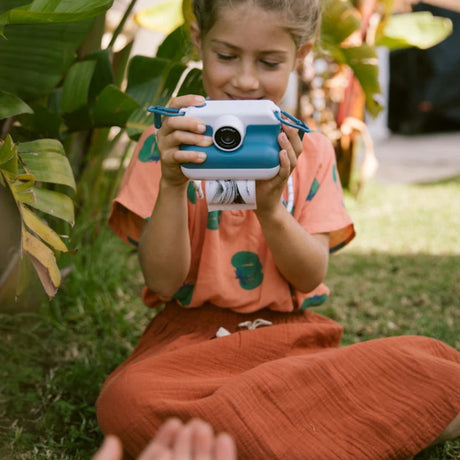 Young girl sitting on grass using a Kidywolf Kidyprint instant camera to print a thermal photo in a garden setting.