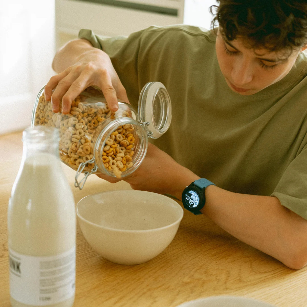 A person pouring round cereal from a glass jar into a white bowl, with a bottle of milk on the table.