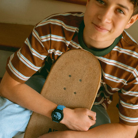 A smiling teen boy holding a skateboard, wearing a striped t-shirt and a smartwatch with a blue strap.
