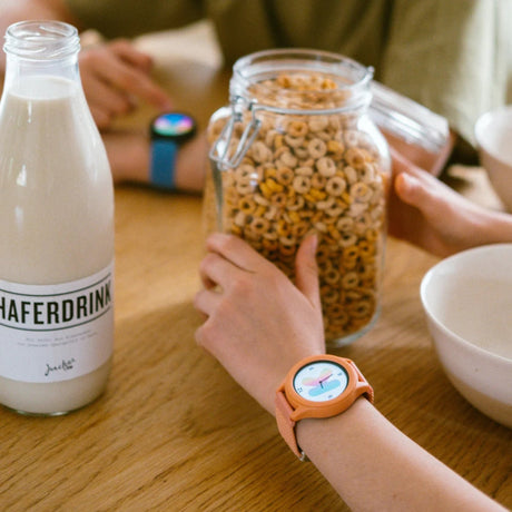 Child wearing a coral smartwatch at a table with cereal and a bottle of oat milk.