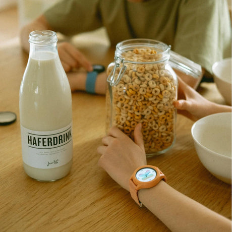 A child wearing a coral smartwatch sitting at a breakfast table with a jar of cereal and oat milk.