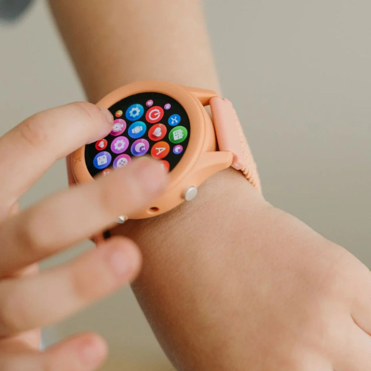 A child's hand tapping the colorful app icons on a peach-colored smart watch screen.