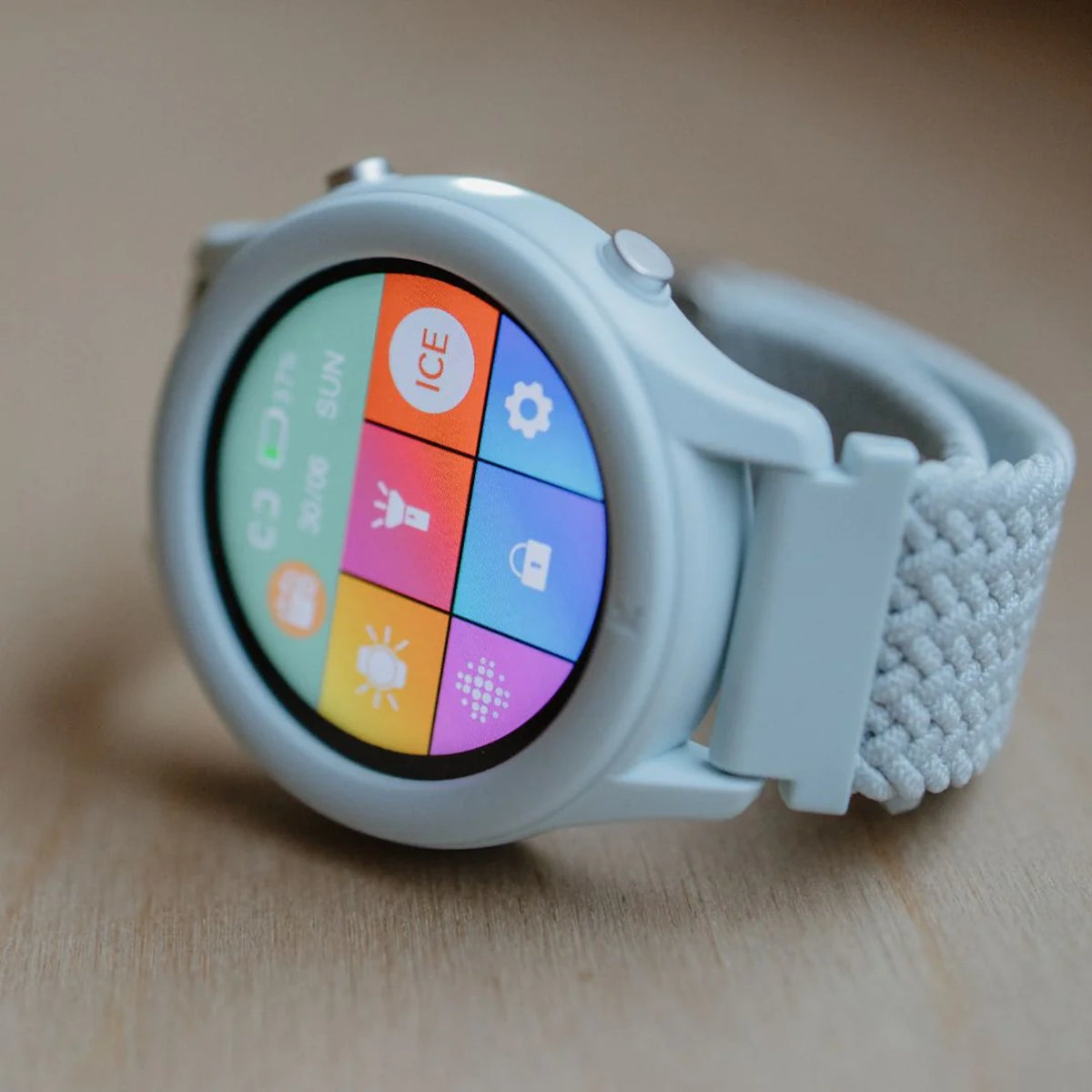 Light blue smartwatch with a colorful grid menu on the screen and a braided strap, resting on a wooden surface.