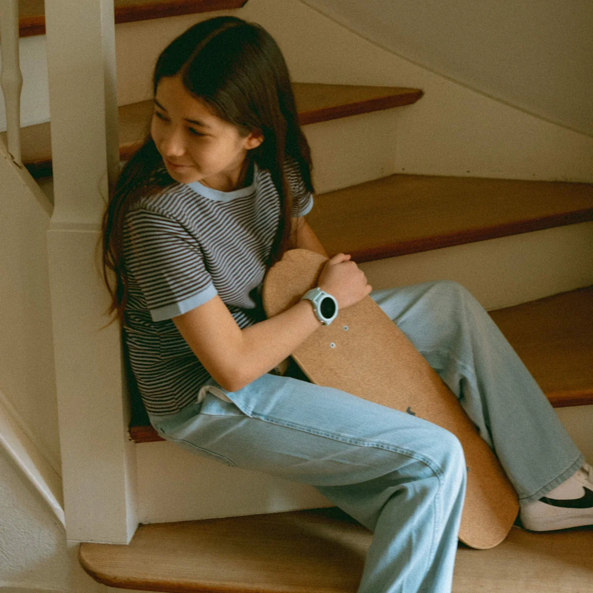 A young girl sits on wooden indoor stairs holding a skateboard and wearing a light blue smartwatch.