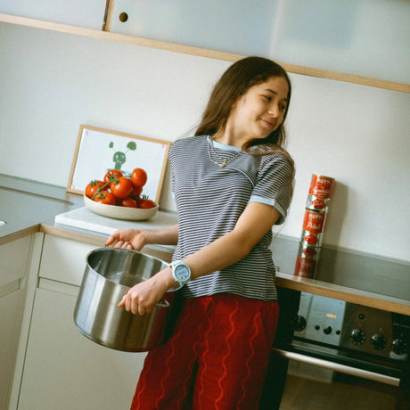 A smiling young girl in a striped shirt holds a large metal cooking pot in a kitchen.