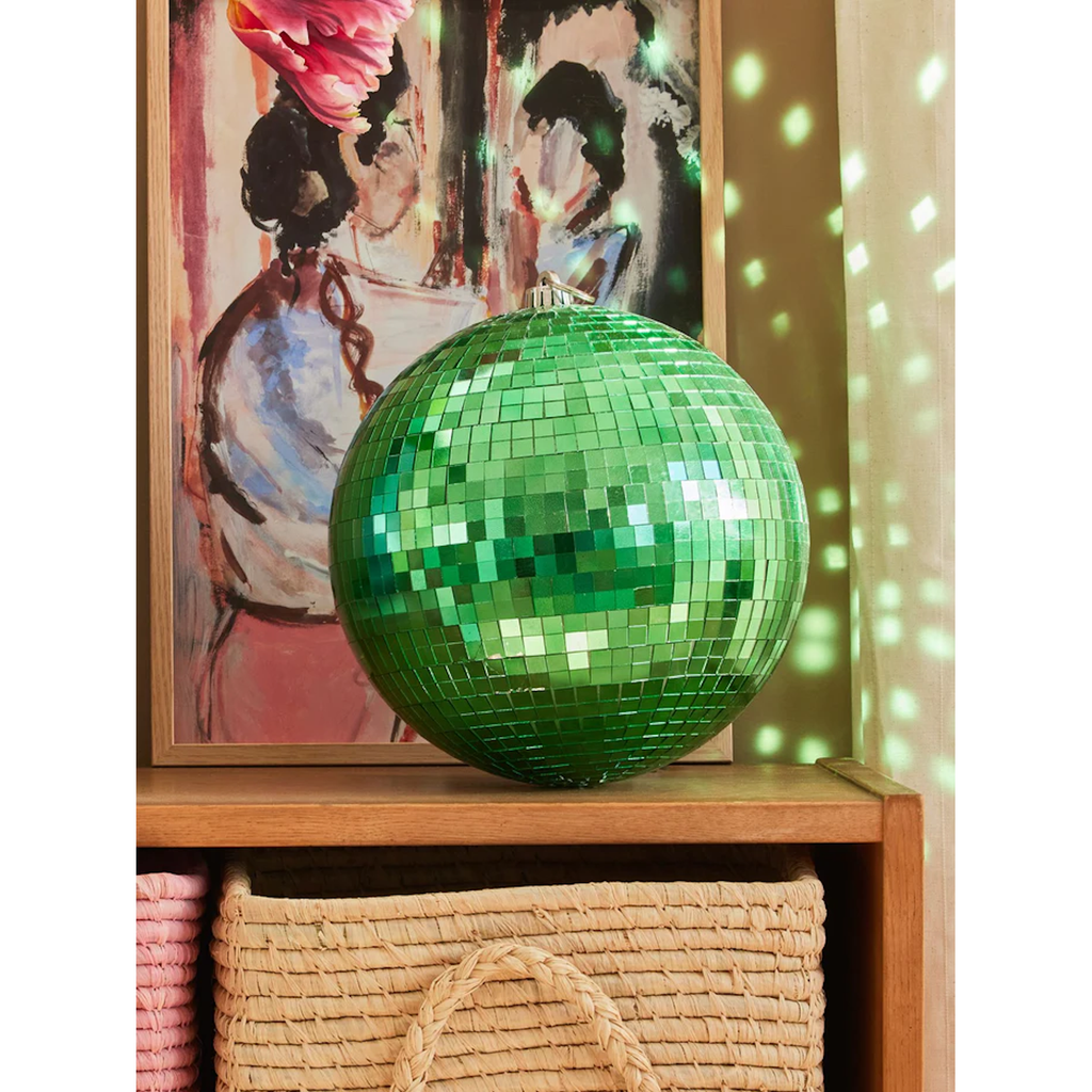 A vibrant green disco ball sits on a shelf, reflecting light onto a painting and a woven basket below. The scene feels artistic and lively.