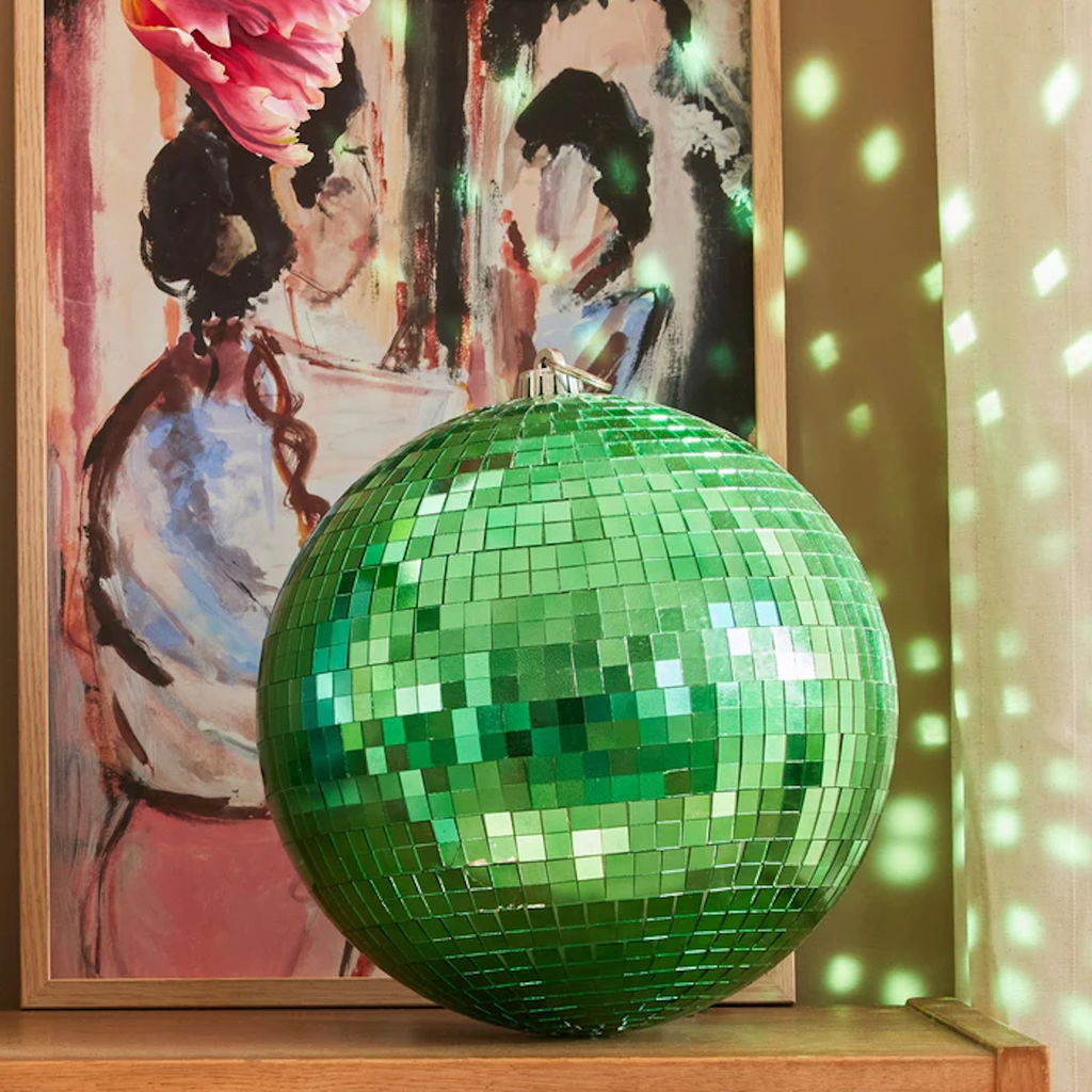 A vibrant green disco ball sits on a shelf, reflecting light onto a painting and a woven basket below. The scene feels artistic and lively.