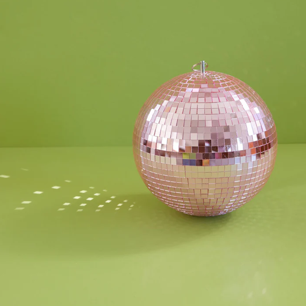 A pink disco ball sits on a light green surface, reflecting scattered light patterns. The scene is bright and playful, evoking a fun, lively atmosphere.