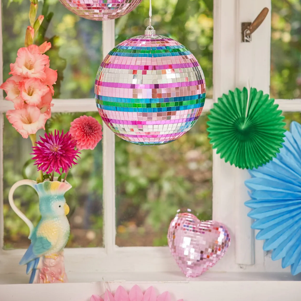 Colorful disco balls, tassels, and paper fans hang by a window with a bird vase and flowers. Vibrant and festive atmosphere.