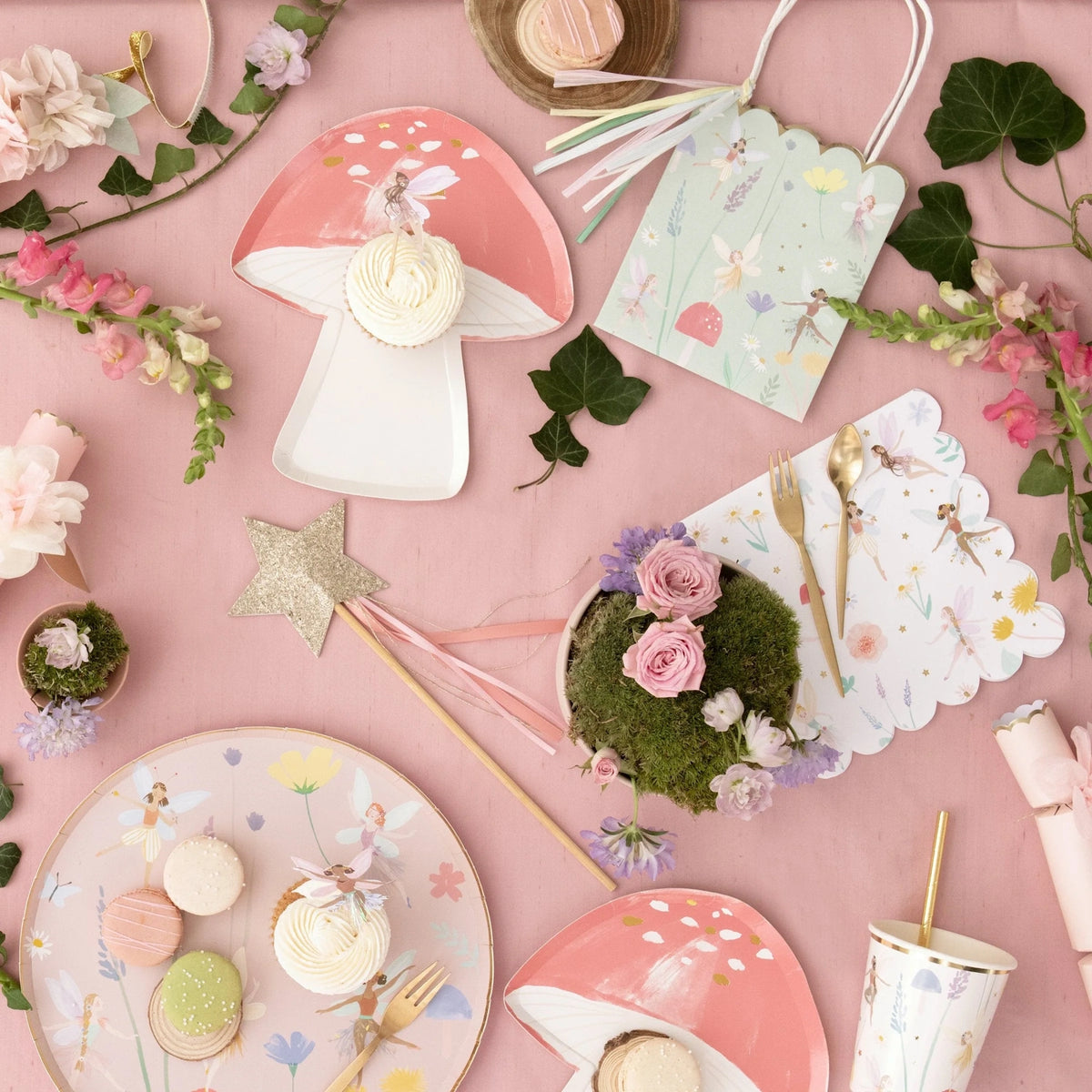 Flat lay of a fairy-themed party table with mushroom plates, cupcakes, macarons, flowers, and a gold wand.