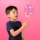 A boy in a navy shirt looks amazed while holding a colorful, glowing balloon against a vibrant pink background. The scene conveys wonder and curiosity.