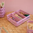 Pink plastic baskets hold colorful spoons and spatulas with floral designs, placed on a woven table. The scene is bright and cheerful.