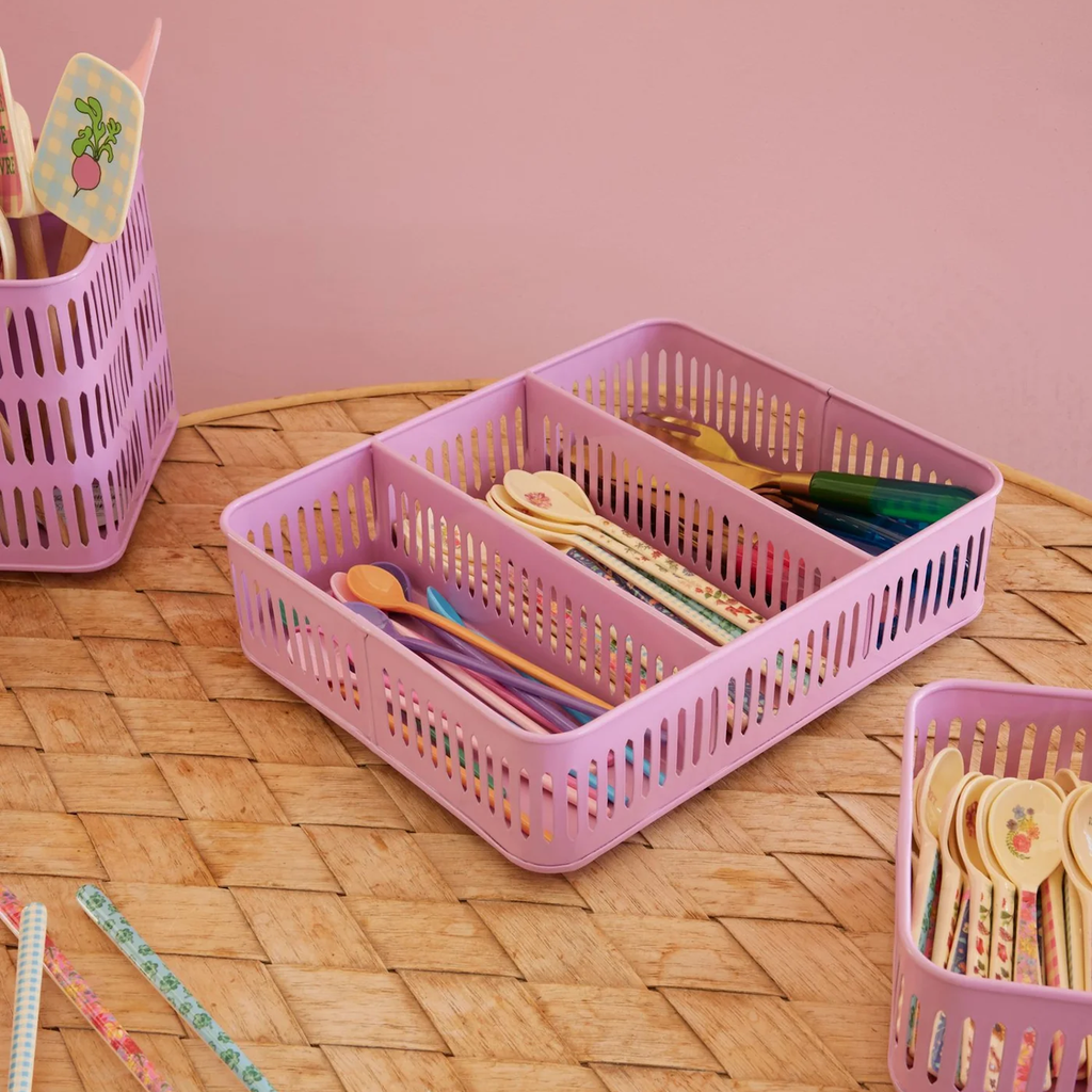 Pink plastic baskets hold colorful spoons and spatulas with floral designs, placed on a woven table. The scene is bright and cheerful.
